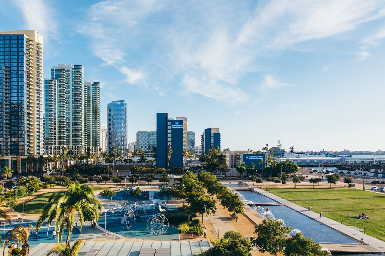 Waterfront Park featuring the playground, interactive water fountains, open green space, and trees set against an urban backdrop of high‑rise buildings